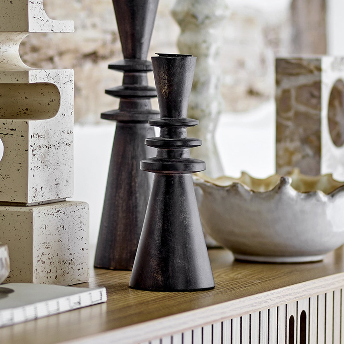Armel Wooden Candle Holder, black mango wood, tall cone base with stacked ring waist, shown among decorative accessories on a wooden sideboard.