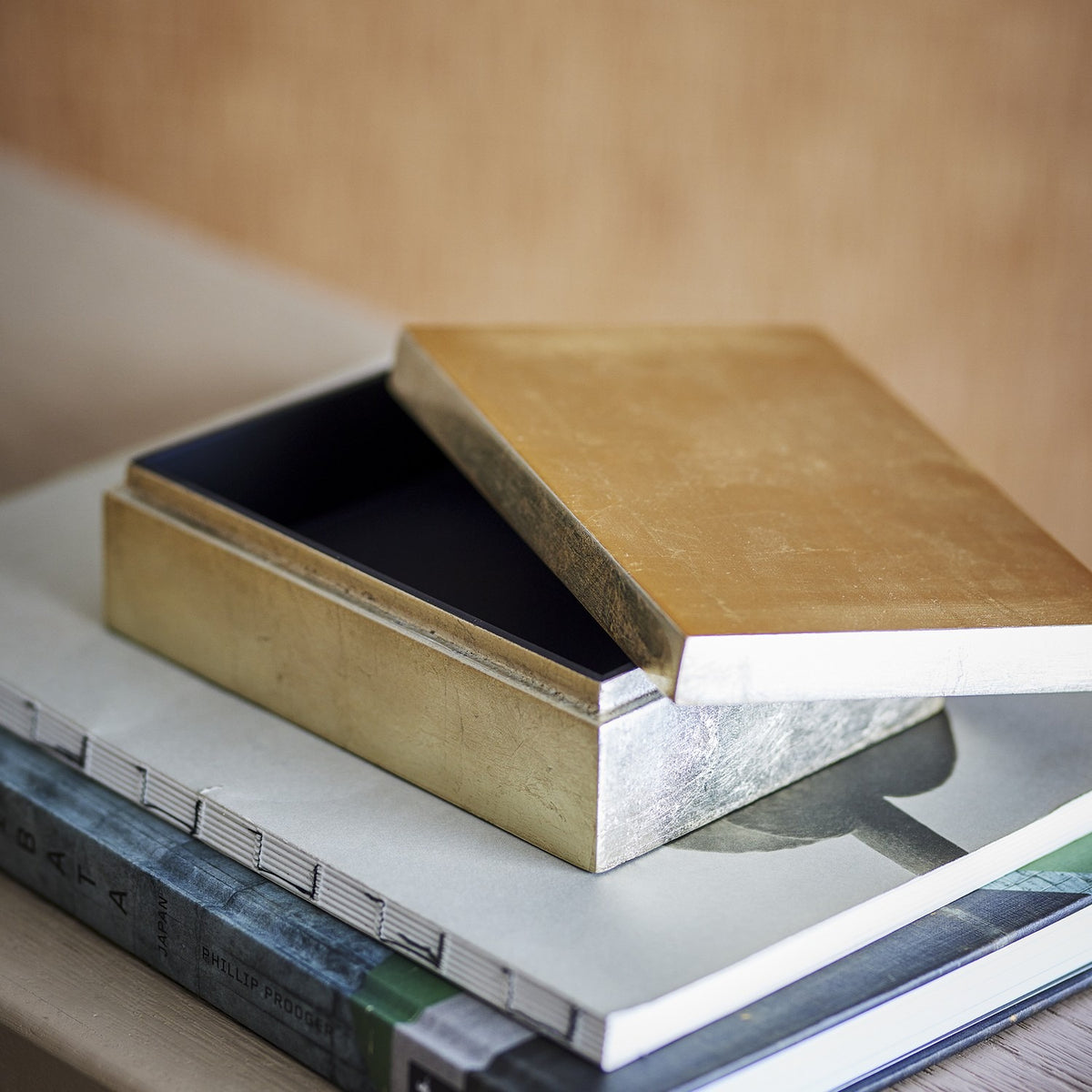 Nenon rectangular storage box in gold MDF with glossy lid ajar revealing a black lacquered interior, shown resting atop stacked books.