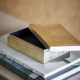Nenon rectangular storage box in gold MDF with glossy lid ajar revealing a black lacquered interior, shown resting atop stacked books.