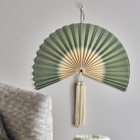 Jaime Bamboo Wall Decor 70cm in Green, a pleated fan-shaped wall hanging with natural bamboo centre and twin tassels, shown mounted above a grey sofa.