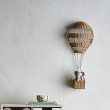 Huggi Rattan Wall Basket in Natural, a hot air balloon-shaped wall decor piece in woven rattan with a hanging basket, mounted on a light grey wall above a shelf with books and a bowl.