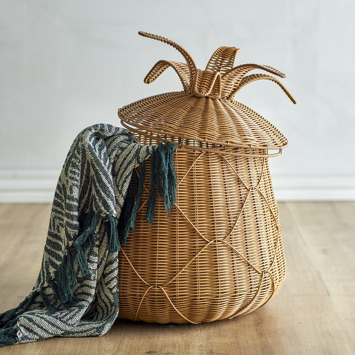 Anann Pineapple Basket with Lid in beige polyrattan, woven with a leafy topped lid, shown beside a striped green throw on a wooden floor.