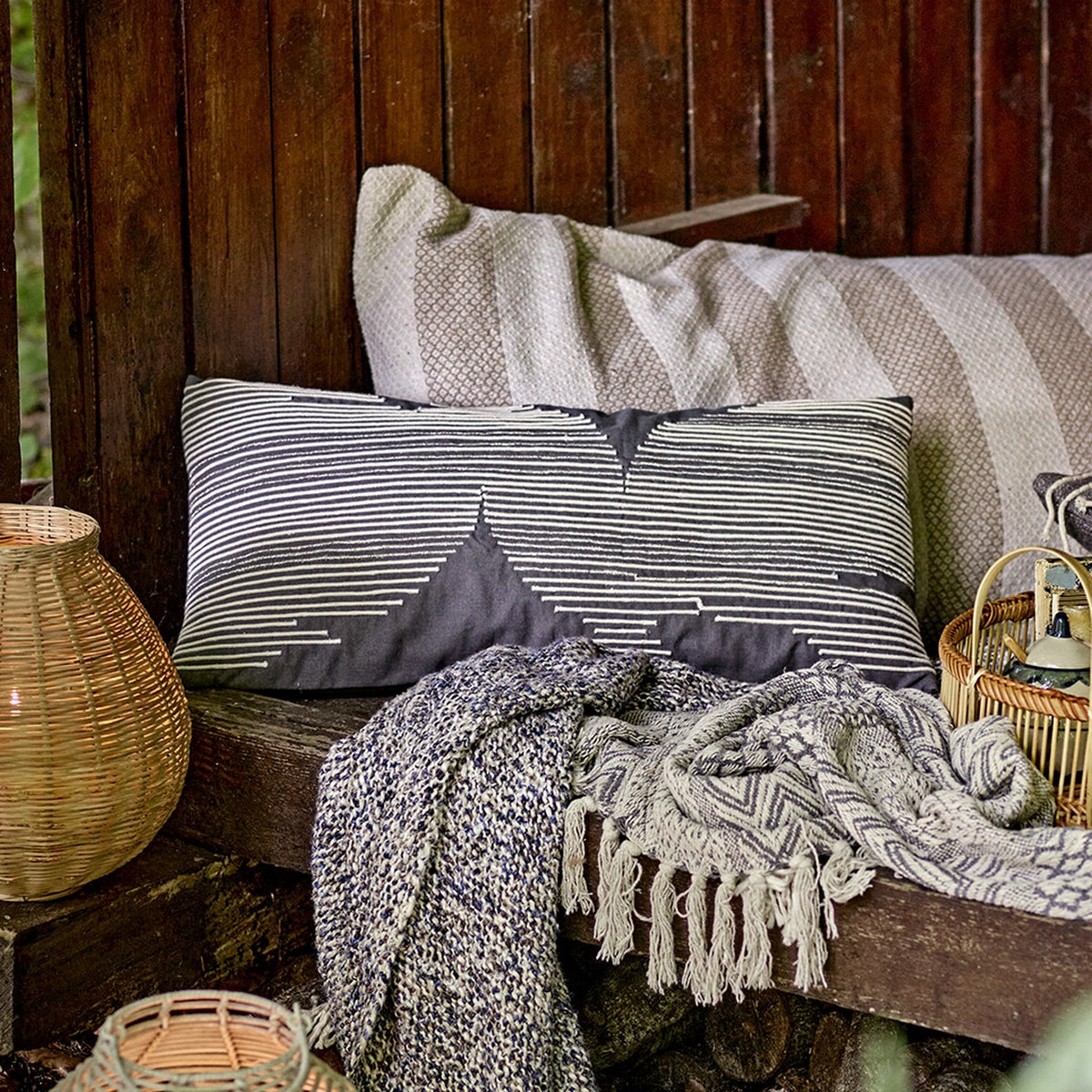 Brechia Striped Cotton Cushion in grey with embroidered stripes forming abstract triangle shapes, styled on an outdoor wooden bench with woven baskets and throws.