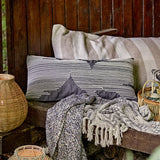 Brechia Striped Cotton Cushion in grey with embroidered stripes forming abstract triangle shapes, styled on an outdoor wooden bench with woven baskets and throws.