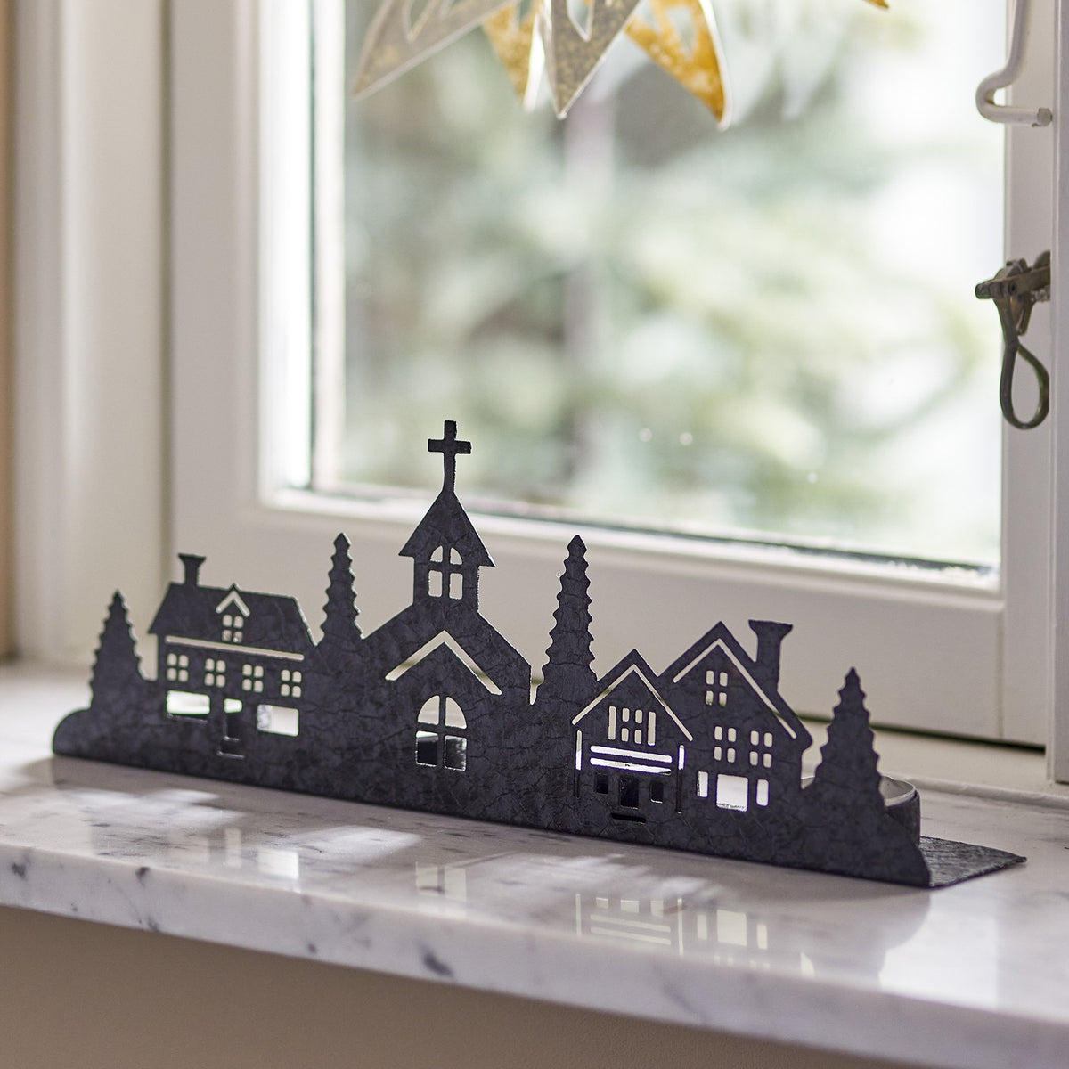 Cielo black iron tealight holder with Christmas village silhouette of houses, fir trees and a church, shown on a marble windowsill.