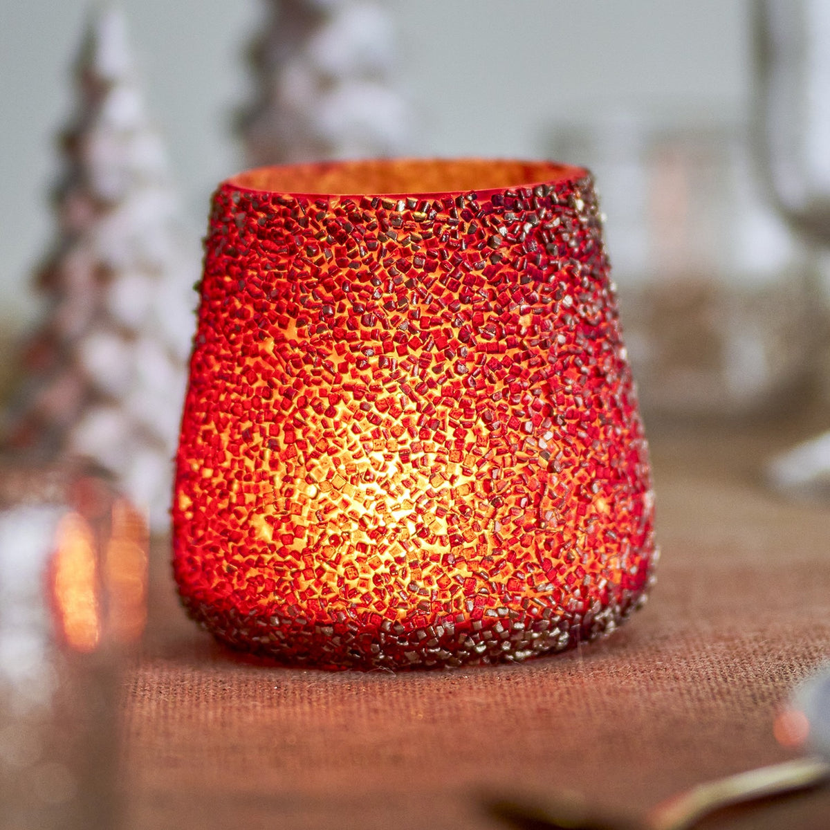 Filiz glass Christmas tealight holder in gold, tapered round shape with dense raised mosaic texture, lit candle glowing inside near blurred Christmas trees.