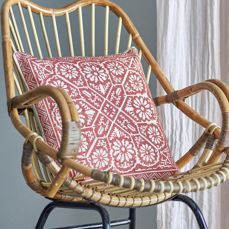 Madita Floral Cotton Cushion in red, square with woven geometric floral pattern, shown on a rattan chair against a grey wall with sheer curtains.