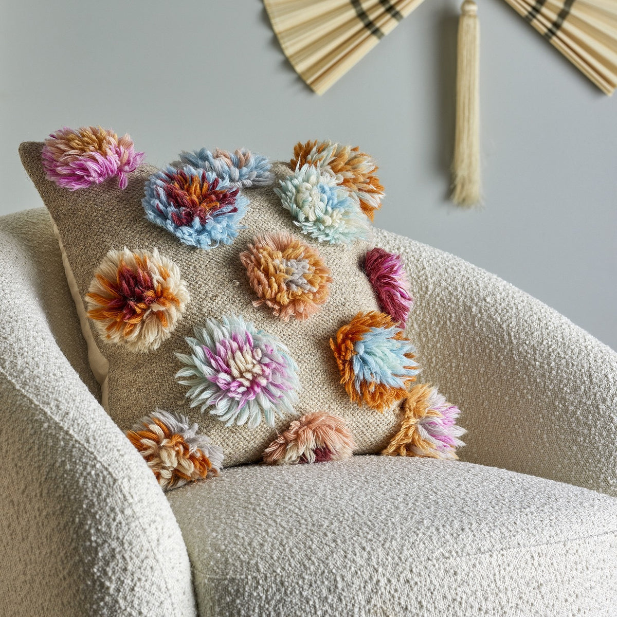 Somma Wool Cushion in Multi with tufted, flower-like yarn clusters on a woven beige base, shown on a bouclé armchair in a living room setting.