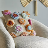 Somma Wool Cushion in Multi with tufted, flower-like yarn clusters on a woven beige base, shown on a bouclé armchair in a living room setting.