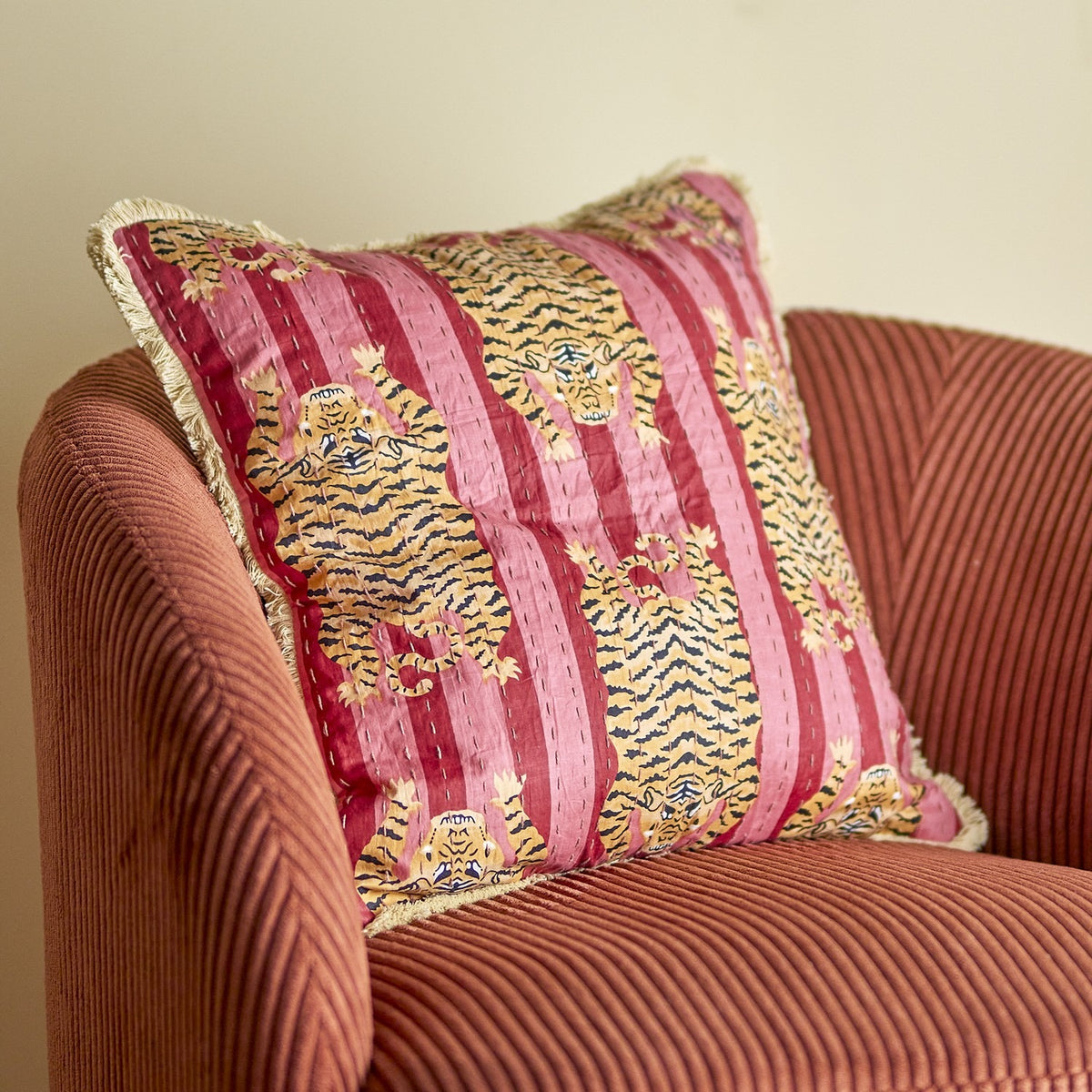 Poirino Cotton Cushion in pink with striped tiger print, fringed edges and visible running stitch detail, shown on a rust corduroy armchair.