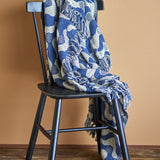 Santena Wavy Cotton Throw in blue with woven wave pattern and fringed edges, draped over a black wooden chair against a warm terracotta wall.
