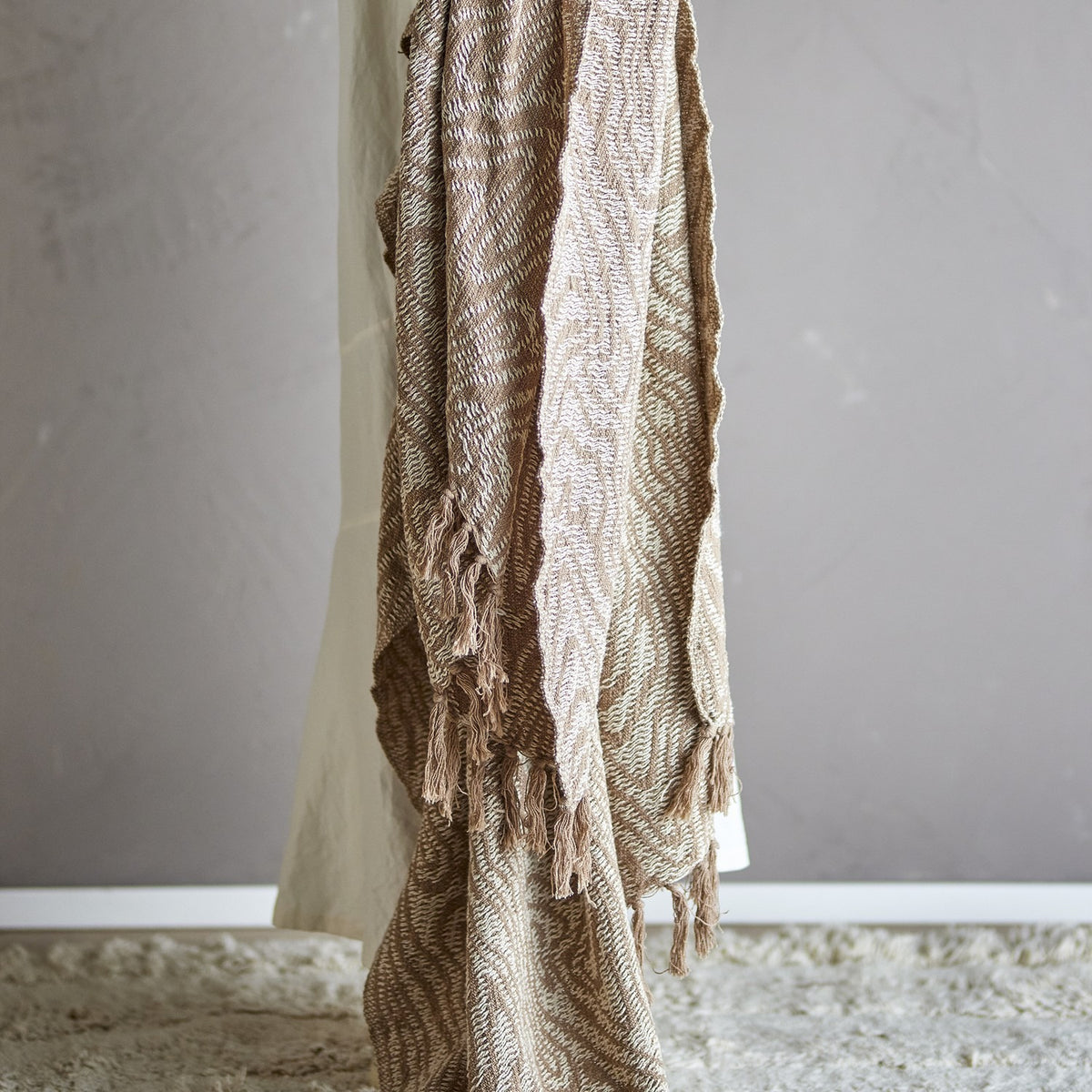 Okapi Cotton Throw in beige with woven wavy-line pattern and fringed edges, draped over a chair against a plain grey wall.