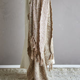 Okapi Cotton Throw in beige with woven wavy-line pattern and fringed edges, draped over a chair against a plain grey wall.
