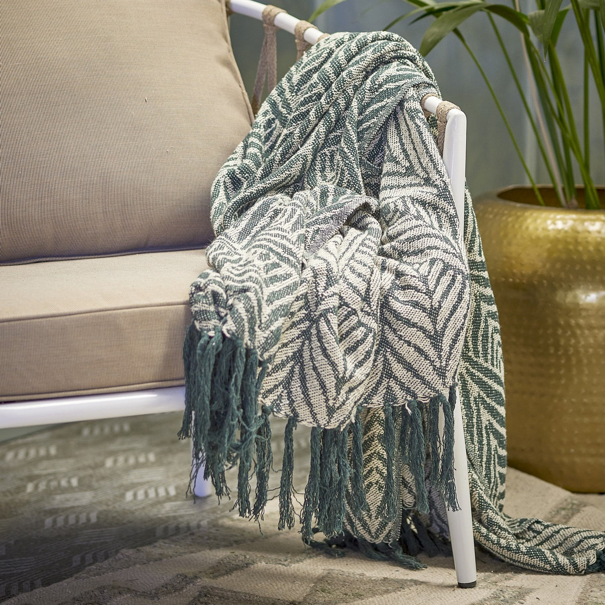 Ugento Leaf Cotton Throw in green, woven with a bold leaf pattern and knotted fringe, draped over a white outdoor sofa frame beside a plant pot.