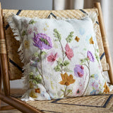 Pogni Floral Cotton Cushion in Natural, a square fringed-edge cover with raised embroidered wildflowers in purple, pink, orange and green, shown on a woven rattan chair.