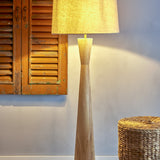Leonor Wooden Floor Lamp with Linen Shade in natural rubberwood, tapered hourglass column and tapered drum shade, shown lit beside rattan stool and wooden shutters.