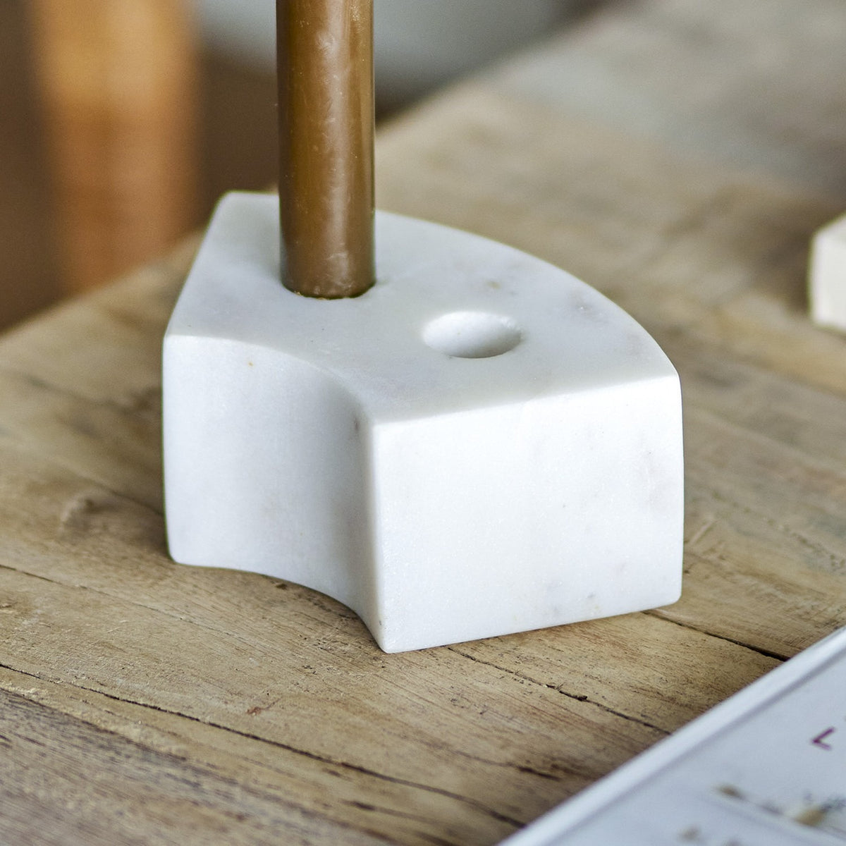 Florida Marble Candle Holder in white, a curved low block with two candle holes, shown holding a brass candlestick on a wooden table.