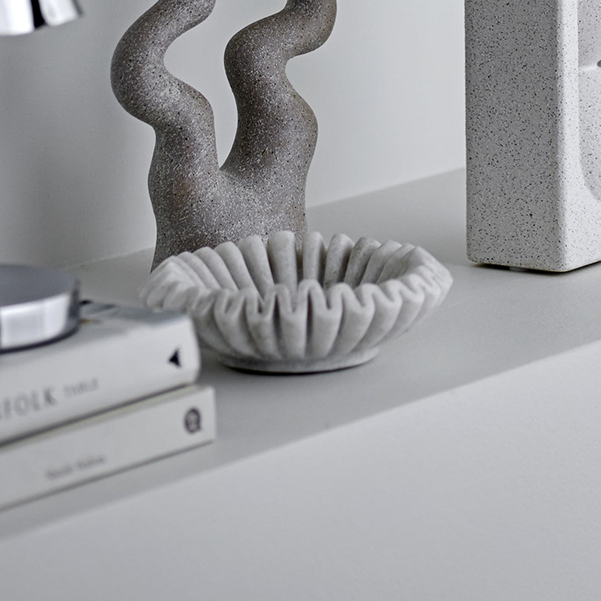 Harmonia Marble Bowl 17.5cm in white with ridged, scalloped petal-like rim, shown on a shelf beside a textured grey sculpture and stacked books.
