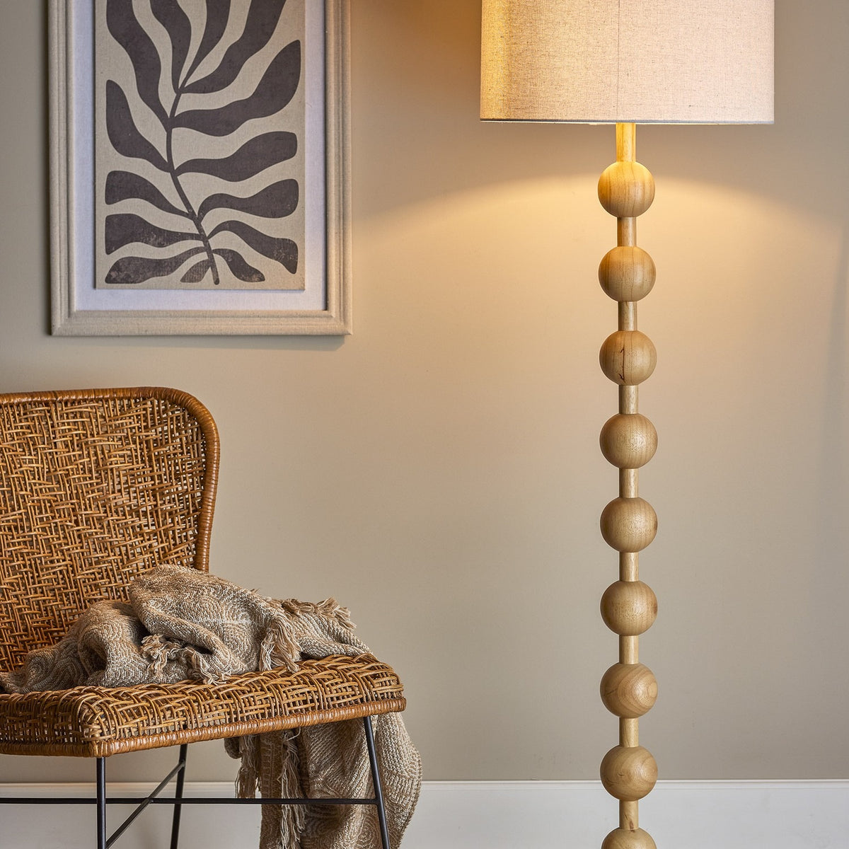 Giacoma Wooden Floor Lamp in natural wood, featuring stacked spherical beads on a slim stem with a linen drum shade, shown lit beside a rattan chair in a living room setting.