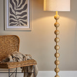 Giacoma Wooden Floor Lamp in natural wood, featuring stacked spherical beads on a slim stem with a linen drum shade, shown lit beside a rattan chair in a living room setting.
