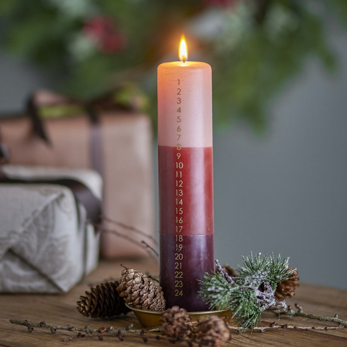 Lit Dip Dye Christmas Candle, a tall pillar in blush, red, and plum ombre wax with a printed 1–24 countdown, displayed on a brass dish with pinecones and festive greenery.