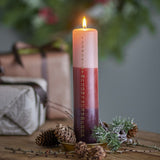 Lit Dip Dye Christmas Candle, a tall pillar in blush, red, and plum ombre wax with a printed 1–24 countdown, displayed on a brass dish with pinecones and festive greenery.
