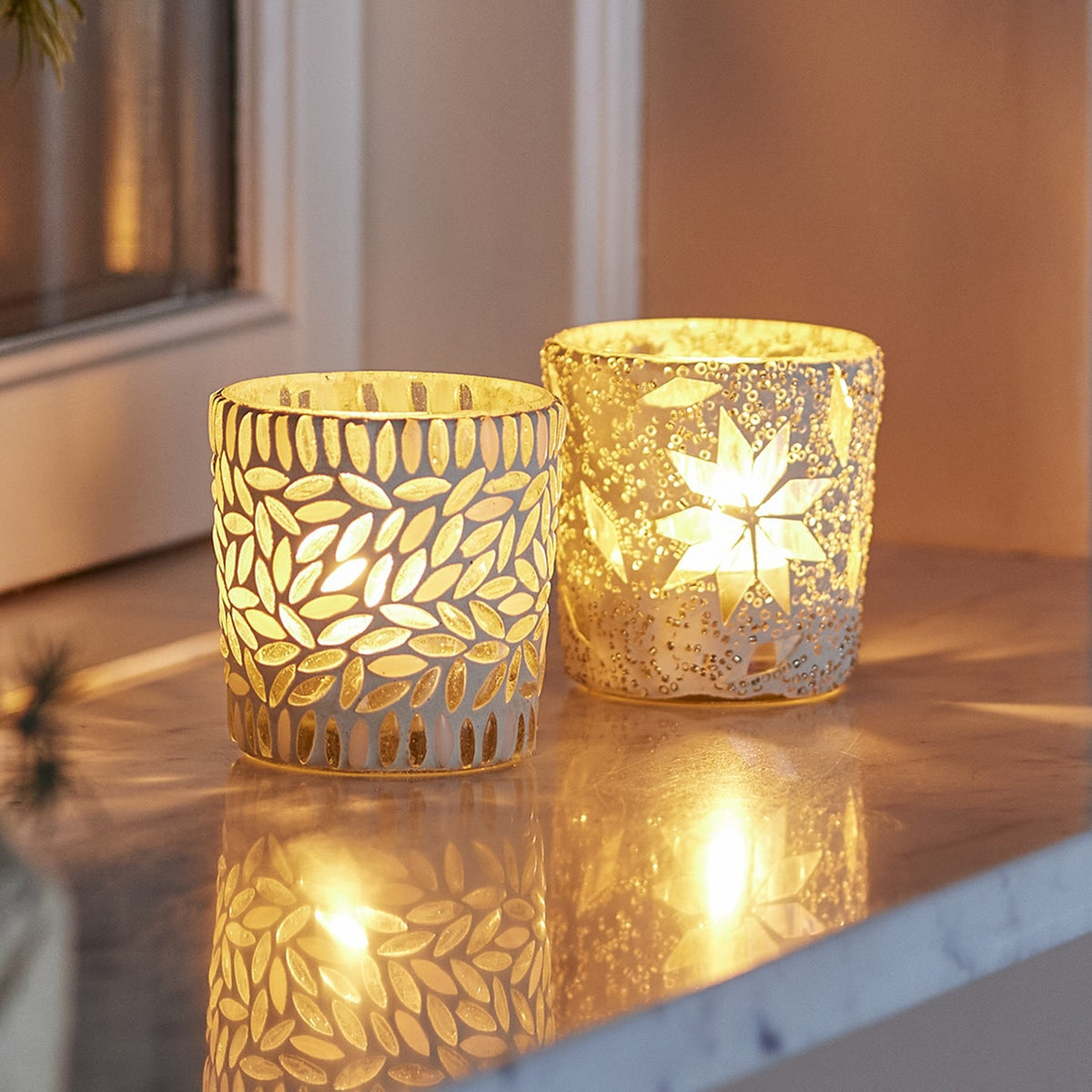 Myr glass tealight holders, set of 2 in white, one with a leaf mosaic design and one with a beaded star pattern, shown lit on a marble surface.