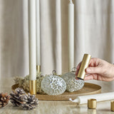 Maxime Iron Christmas Candle Holders Set of 7 in gold, round textured tray with slim tube holders and white taper candles, styled with pinecones and a hand placing a holder.