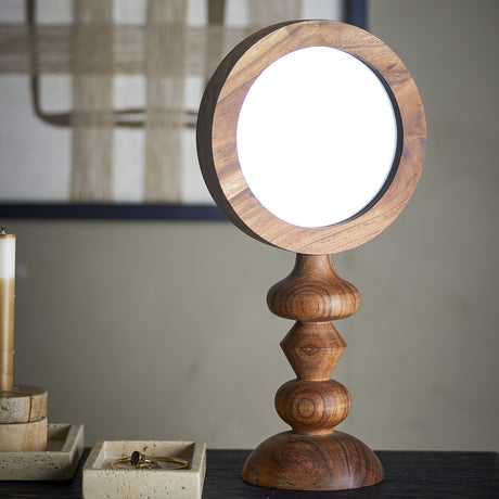 Malukka Wooden Table Mirror in brown acacia wood, round glass face set in a thick frame atop a turned stem with stacked bulb and diamond details, shown on a bedroom dresser.
