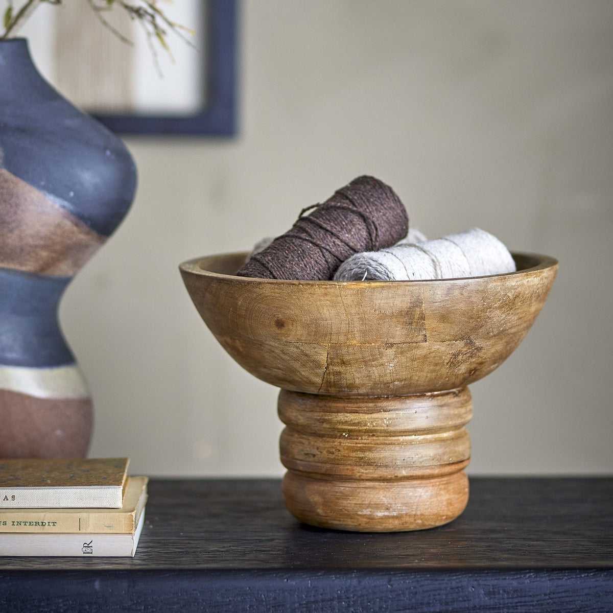 Nanuka round wooden pedestal bowl in brown mango wood with ringed foot, shown holding wound yarn skeins on a dark console in a living room setting.