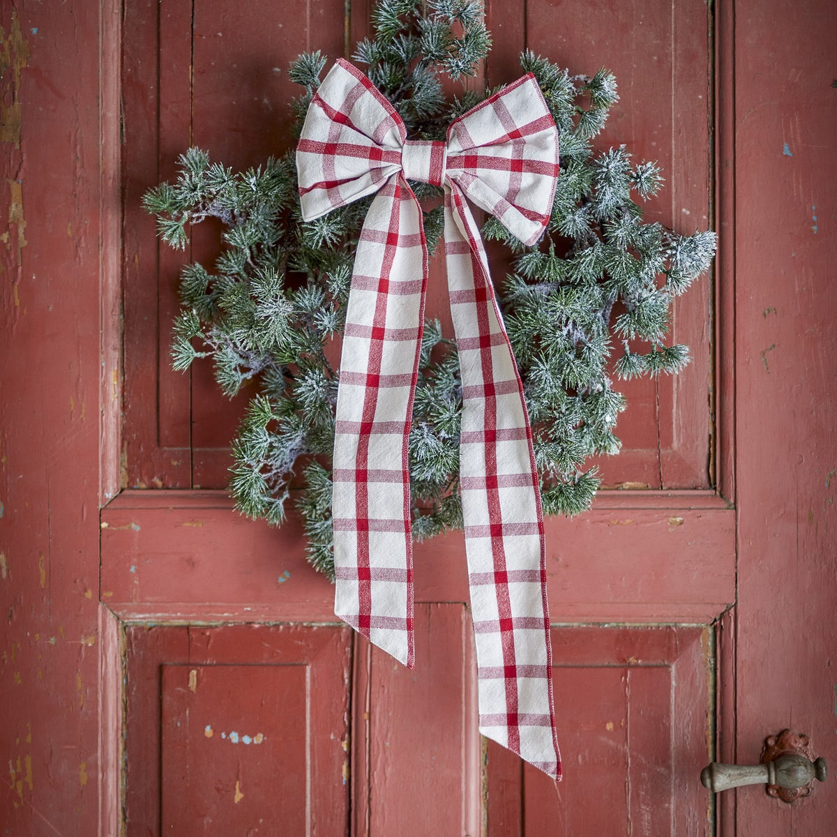 Ruby Cotton Christmas Bow in red check fabric with rounded loops and long tails, fixed to a frosted pine wreath on a weathered red door.