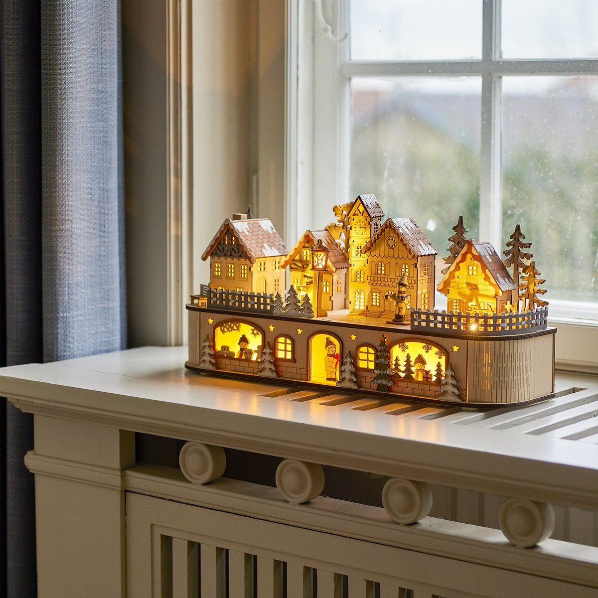 Norwich Wooden Christmas Decoration with Light in beige, a lit-up wooden village with gabled houses, clock tower and fir trees, shown glowing on a windowsill.