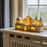 Norwich Wooden Christmas Decoration with Light in beige, a lit-up wooden village with gabled houses, clock tower and fir trees, shown glowing on a windowsill.