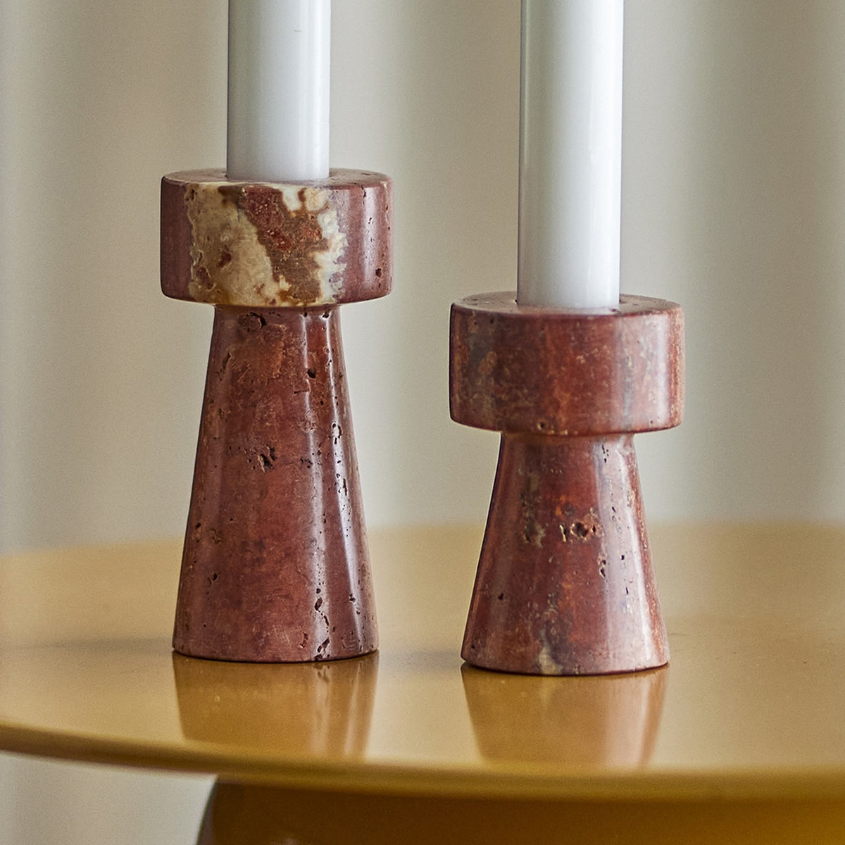Anneli Travertine Candle Holders Set of 2 in Brown, two tapered stone bases with round collars holding white candles, shown at different heights on a glossy mustard table.