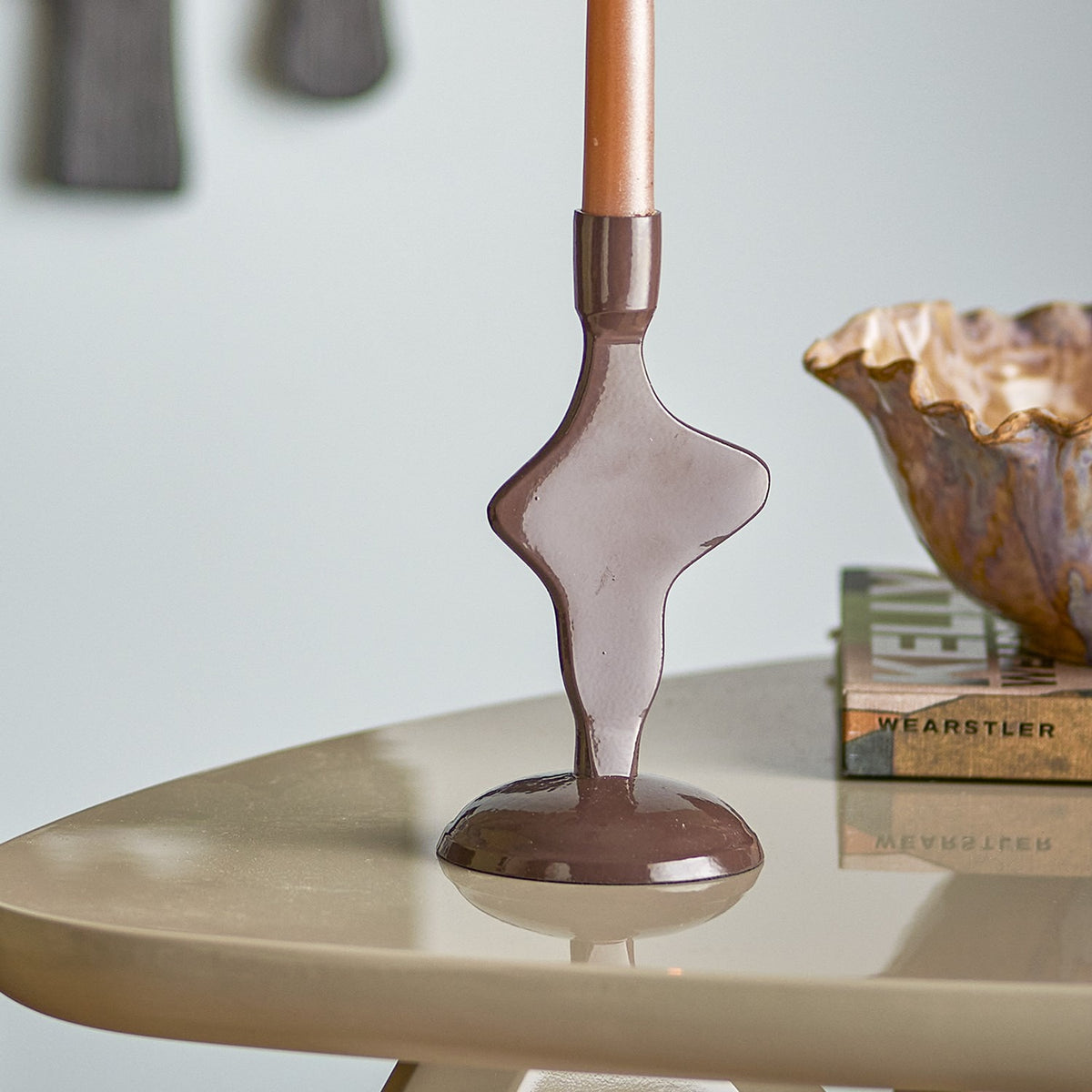 Cidra Aluminium Candle Holder in brown, an abstract sculptural base with glossy powder-coated finish, holding a taper candle on a round table beside a scalloped bowl and books.