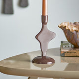 Cidra Aluminium Candle Holder in brown, an abstract sculptural base with glossy powder-coated finish, holding a taper candle on a round table beside a scalloped bowl and books.