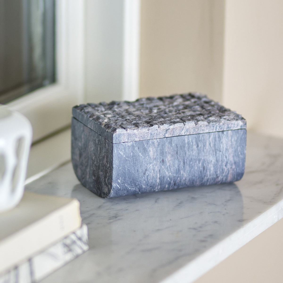 Nubia Soapstone Storage Box with Lid in grey, veined stone with a textured raised-square lid, shown on a marble windowsill beside books and a lamp base.