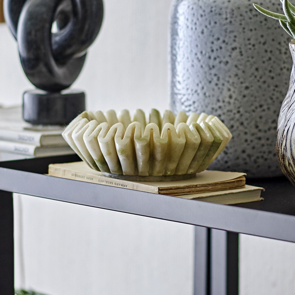 Harmonia Fluted Marble Bowl 17.5cm in white, with deep pleated flutes and a wavy rim, styled on a book atop a dark console table beside vases and a sculpture.