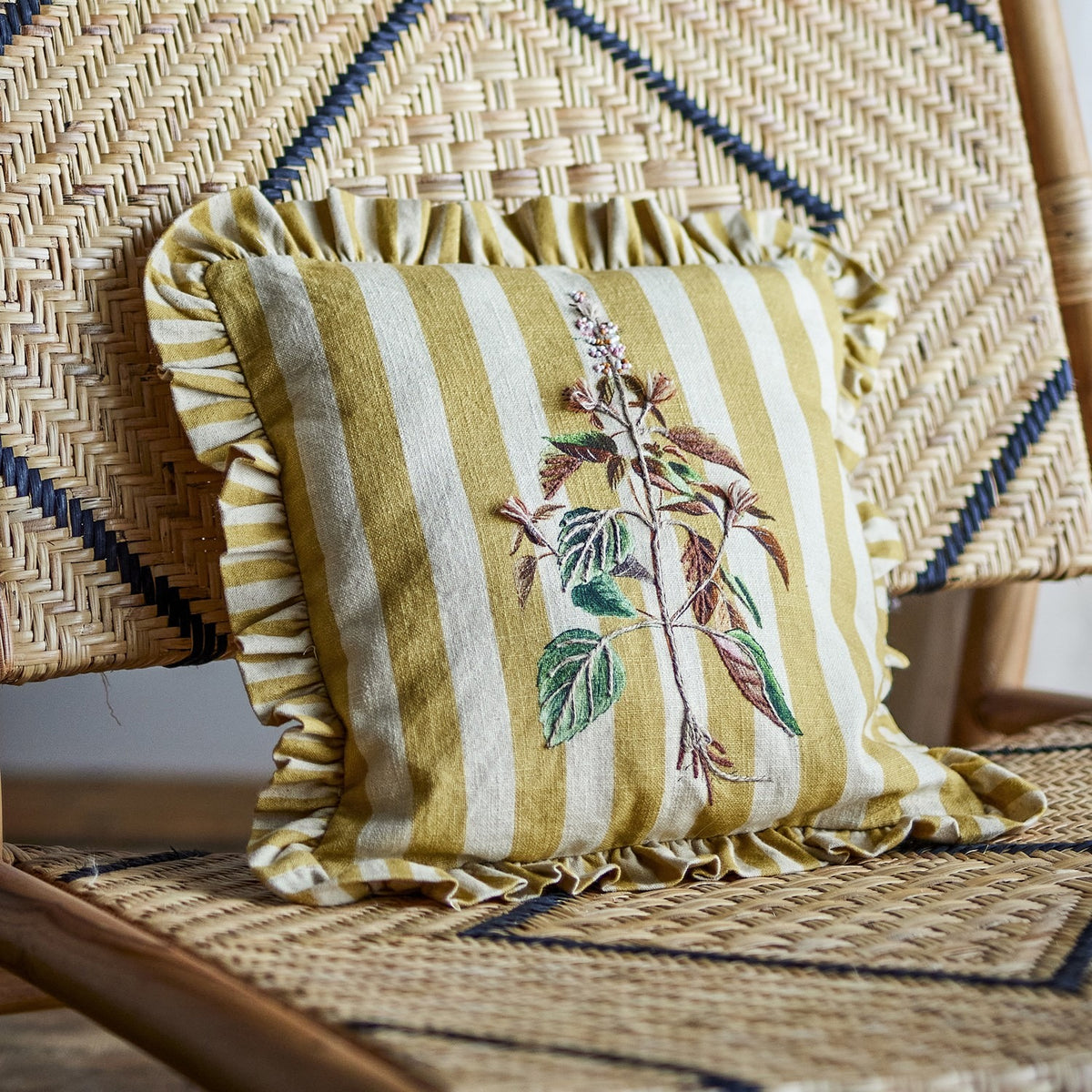 Camila Striped Cotton Cushion in yellow, square with ruffled edge, embroidered botanical stem design, shown on a woven rattan chair.