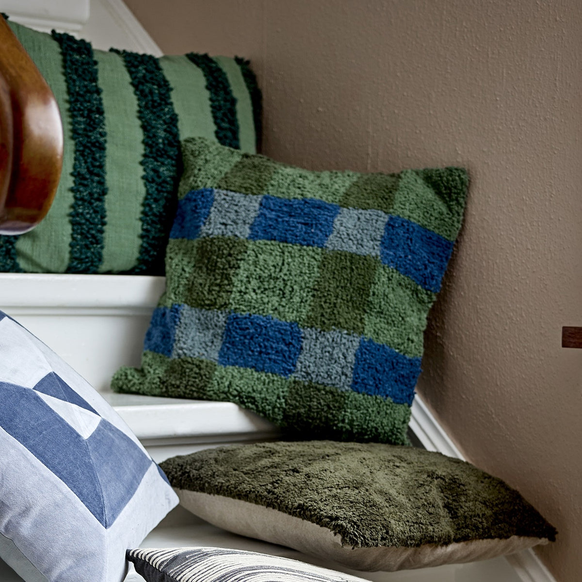 Pil Cotton Cushion in Green, a square tufted cotton cushion with a blue and green check pattern, shown against a plain striped cushion in a cosy corner setting.