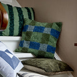 Pil Cotton Cushion in Green, a square tufted cotton cushion with a blue and green check pattern, shown against a plain striped cushion in a cosy corner setting.