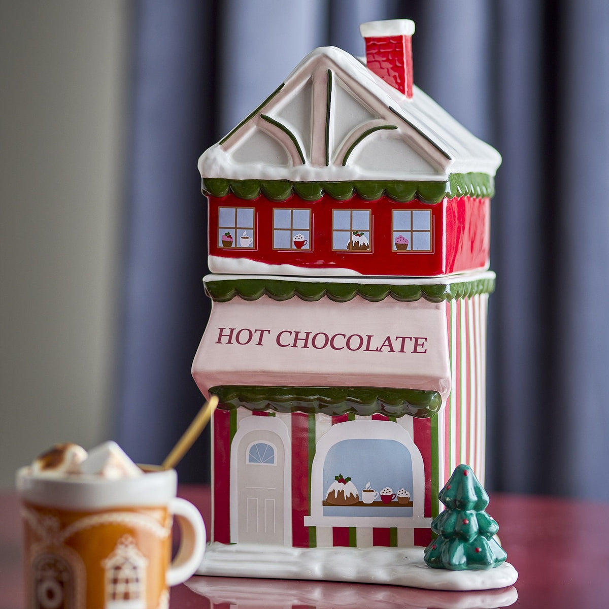 Gloria ceramic Christmas storage jar shaped like a shop, red and white with snow-topped roof, chimney, and "HOT CHOCOLATE" awning, shown beside a mug on a table.