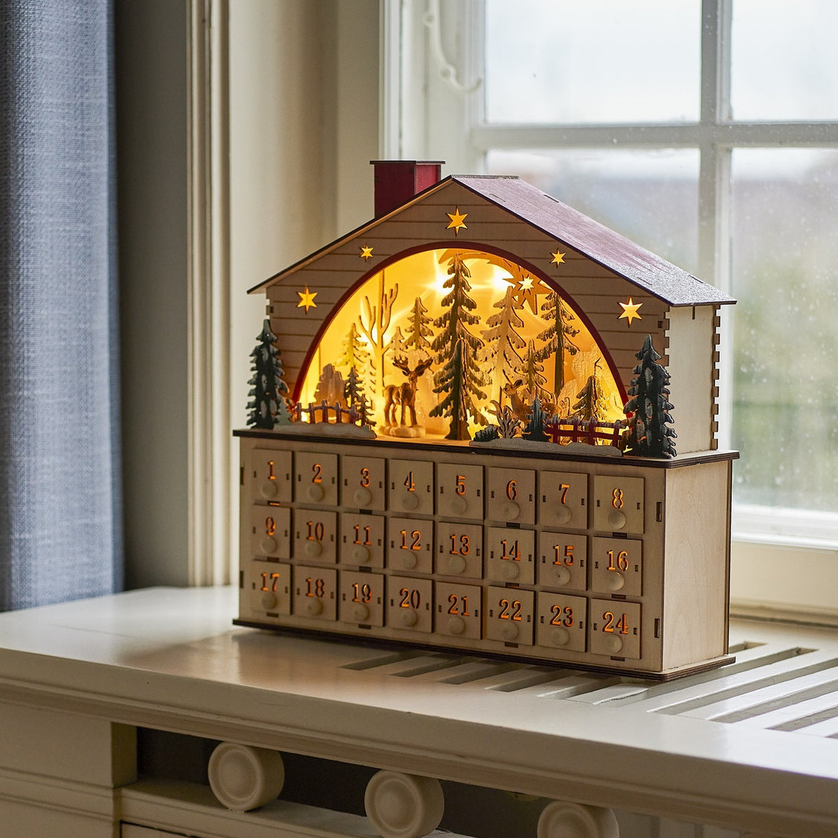 Beige wooden house-shaped advent calendar with pitched roof, chimney, cut-out stars, a lit arched window showing a winter forest scene with a deer, and 24 numbered drawers.