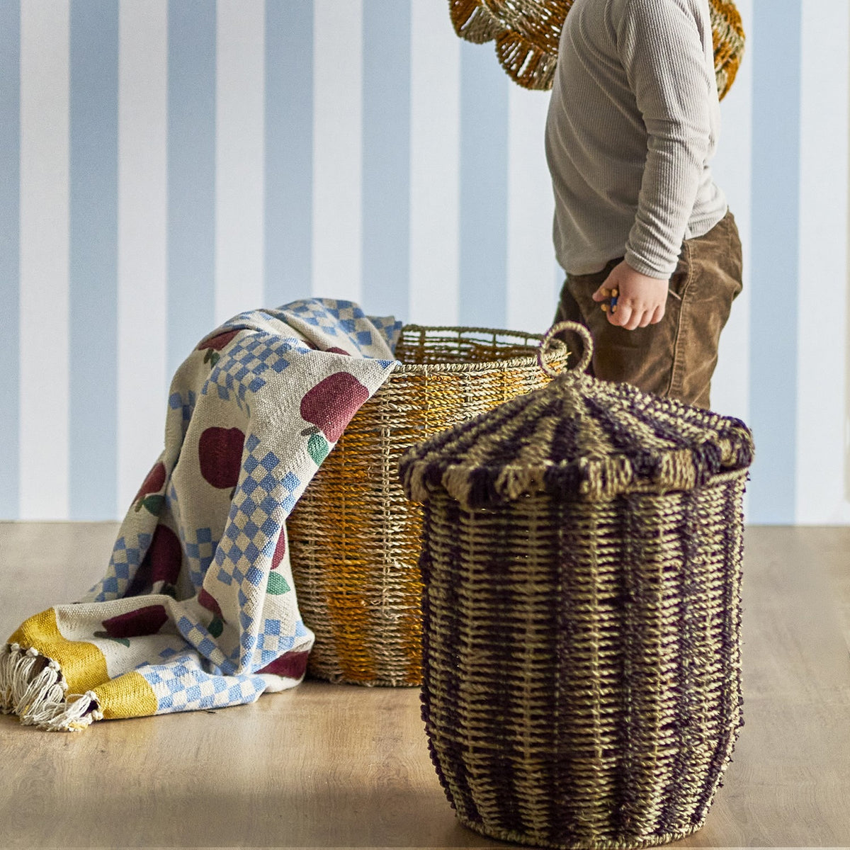Cikkeline striped seagrass lidded storage basket with pointed lid and woven loop handle, shown in multicoloured bands beside a child and folded blanket in a striped-wallpaper room.