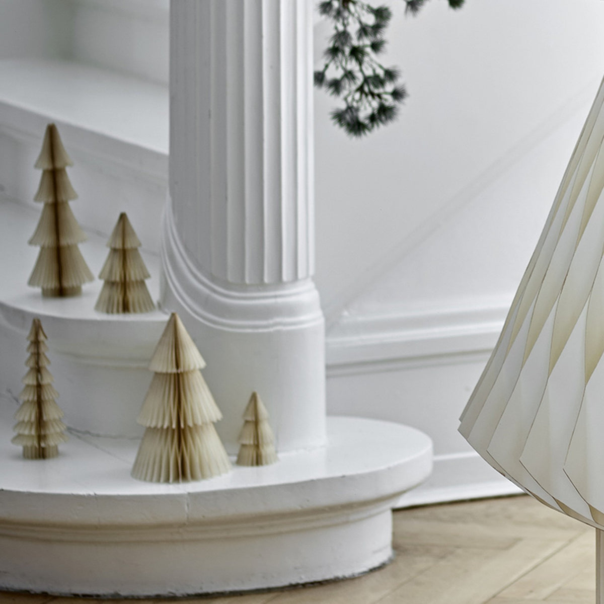 Milan Paper Christmas Decorations Set of 5 in cream, tiered folded-paper trees of varying heights displayed on a white staircase ledge beside a column.