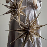 Ilo Paper Christmas Stars Set of 3 in brown, two multi-pointed star decorations with teardrop cut-outs hanging against a person's dress, shown in a lifestyle setting.