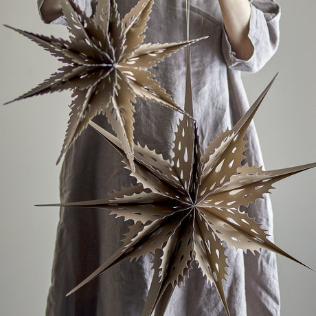 Ilo Paper Christmas Stars Set of 3 in brown, two multi-pointed star decorations with teardrop cut-outs hanging against a person's dress, shown in a lifestyle setting.