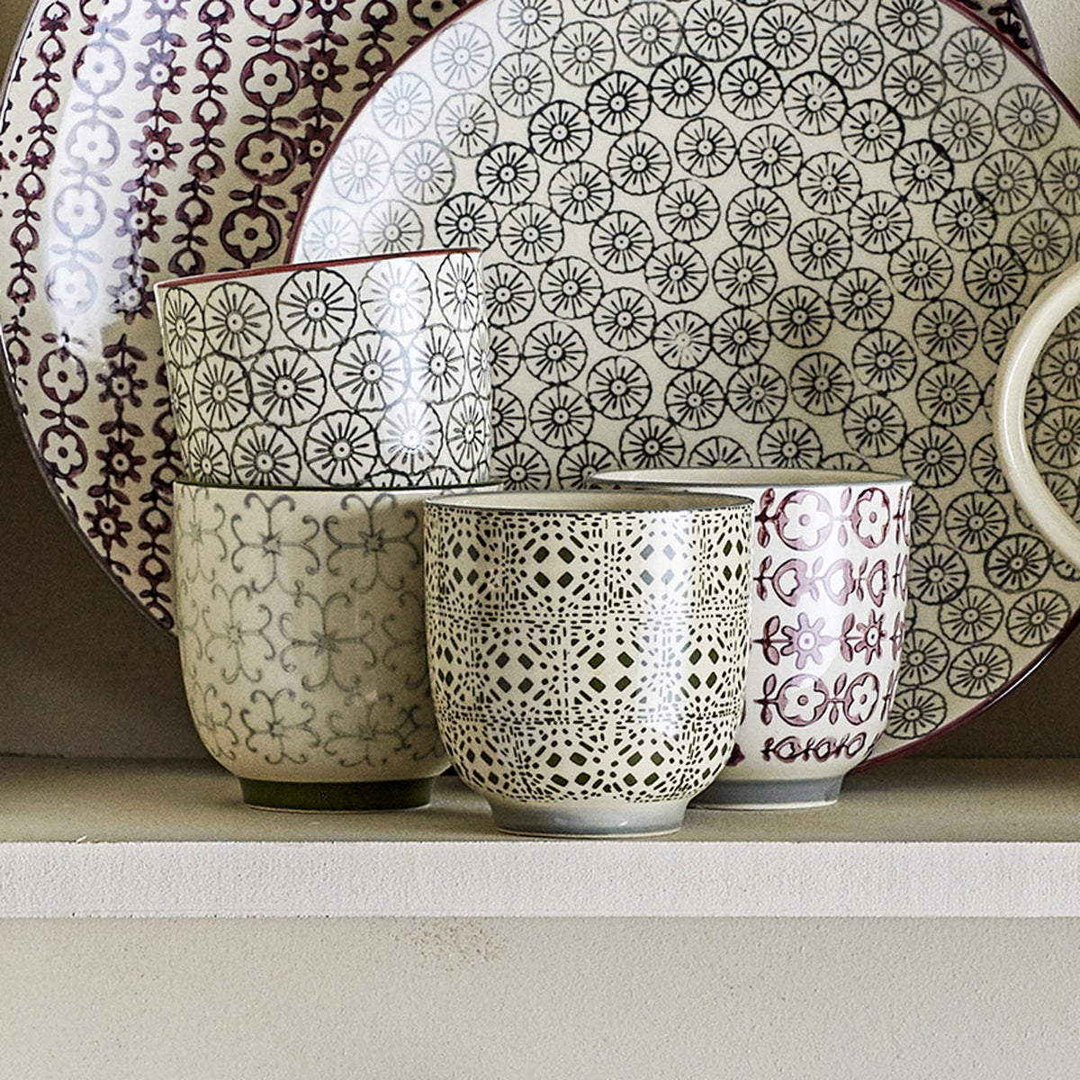 Karine Floral Stoneware Cups, set of four handleless 150ml cups, each with a different hand-stamped floral or geometric pattern, shown on a shelf with patterned plates.