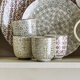 Karine Floral Stoneware Cups, set of four handleless 150ml cups, each with a different hand-stamped floral or geometric pattern, shown on a shelf with patterned plates.