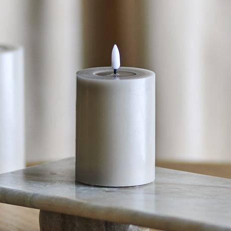 Ponza LED Candle in grey, a paraffin pillar candle with a flame-shaped tip set in a shallow top dip, shown on a marble surface with a blurred curtain backdrop.